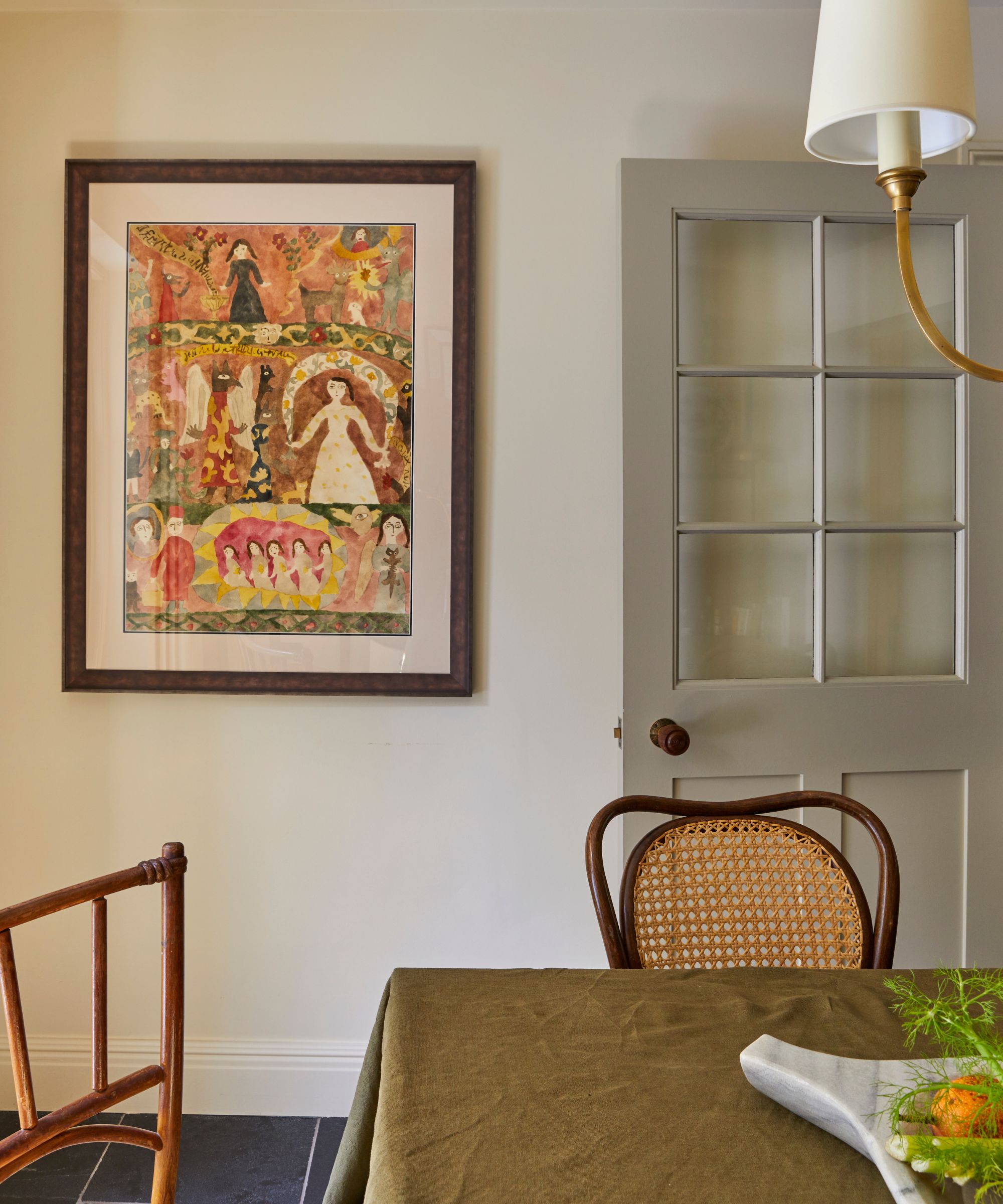 Dining table with olive green tablecloth, vintage wooden chairs, folk art on the wall, a marble fruit bowl and grey painted door