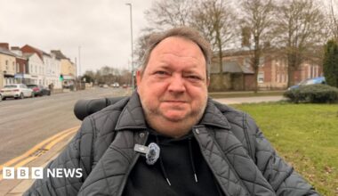 A man sitting in a wheelchair on a path looking at the camera. He is wearing a black hoodie and a black padded jacket. He has short brown hair and stubble. Large buildings can be seen on either side of a road, with cars parked by the side of the road.