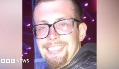 A man with short brown hair and a short brown goatee. He is wearing black framed glasses and a black leather jacket.