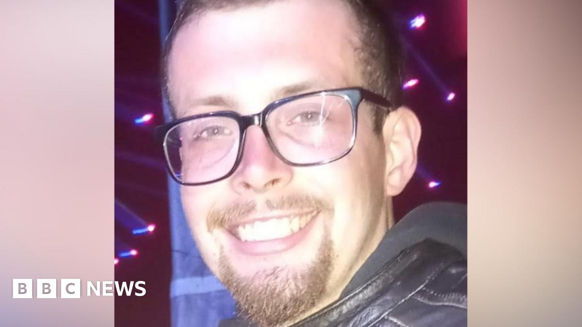 A man with short brown hair and a short brown goatee. He is wearing black framed glasses and a black leather jacket.