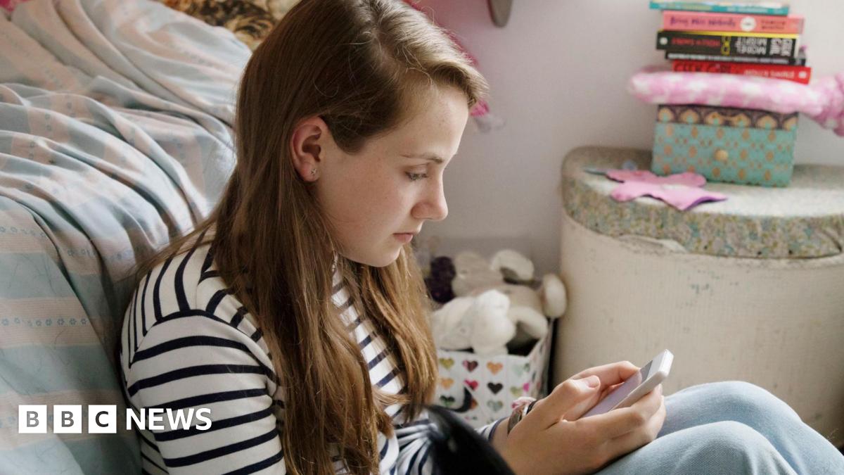 A teenage girl with long-brown hair leans against her bed as she sits on the floor and looks at her phone