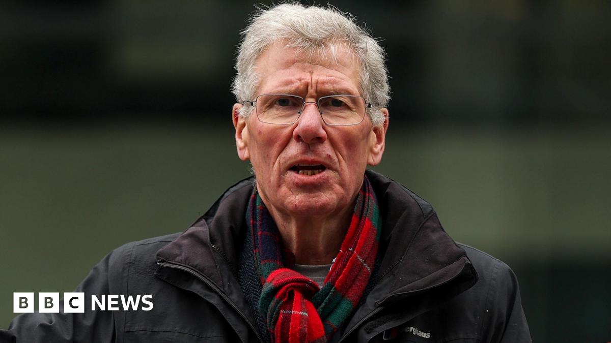 Kenny MacAskill has short grey hair and wears silver wire rimmed glasses. He wears a red and green tartan scarf and black rain jacket and looks directly into the camera