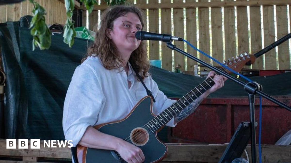 Ben Smith, who has shoulder-length hair and is wearing a white shirt, playing a guitar and singing into a microphone. He is smiling and looking into the distance.
