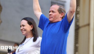 Juan Pablo Guanipa (right) holds his arms in the air while wearing a blue t-shirt beside Maria Corina Machado (left), wearing a white t-shirt and holding a microphone.