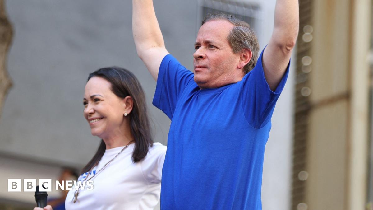 Juan Pablo Guanipa (right) holds his arms in the air while wearing a blue t-shirt beside Maria Corina Machado (left), wearing a white t-shirt and holding a microphone.