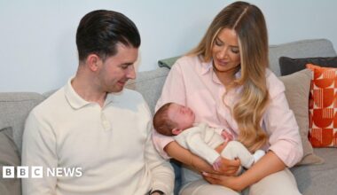 A young couple sit on a sofa. The woman cradles a newborn baby in her arms.
