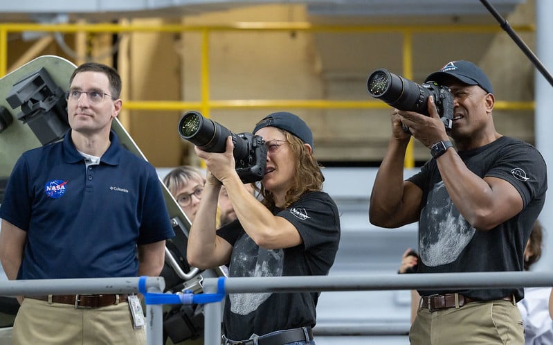 Three people stand indoors, two holding large cameras and taking photos. The person on the left wears a NASA shirt and glasses, while the other two wear matching t-shirts and baseball caps. Industrial equipment is visible in the background.