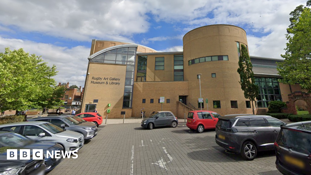 A modern, light-coloured brick building with lettering on the side which indicates it houses an art gallery, museum and library. There is a car park in the foreground of the image with car parks in rows on either side. There are a number of trees nearby the building.