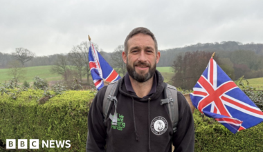 A man with dark hair and a dark beard wearing a black hoodie wears a grey rucksack on his back with two union jack flags coming from his back. He is stood in the countryside with a large hedge behind him.