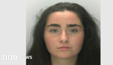 A police mugshot of Liberty Mitchell. She has thick eyebrows, long dark hair and grey-blue eyes, and is looking right at the camera with a blank expression.