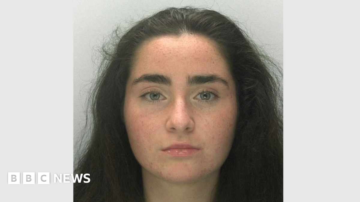 A police mugshot of Liberty Mitchell. She has thick eyebrows, long dark hair and grey-blue eyes, and is looking right at the camera with a blank expression.