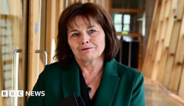 Jeane Freeman in green dress jacket, walks along a corridor in the Scottish Parliament. She has medium-length dark hair.