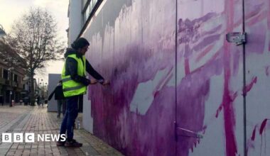 A person standing on a street wearing a high vis jacket painting a wall with purple paint.