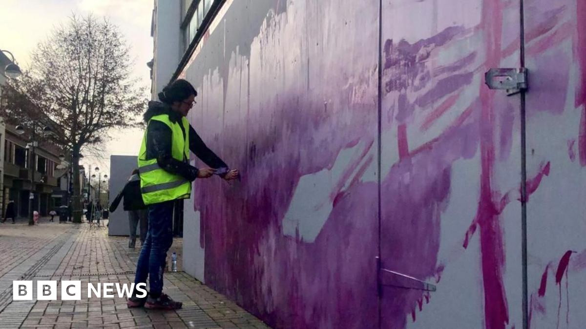A person standing on a street wearing a high vis jacket painting a wall with purple paint.
