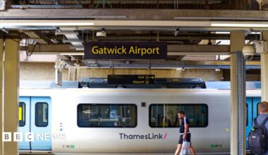 Sign at Gatwick Airport railway station with platform and a white Thameslink train departing the station. People are stood on the platform.
