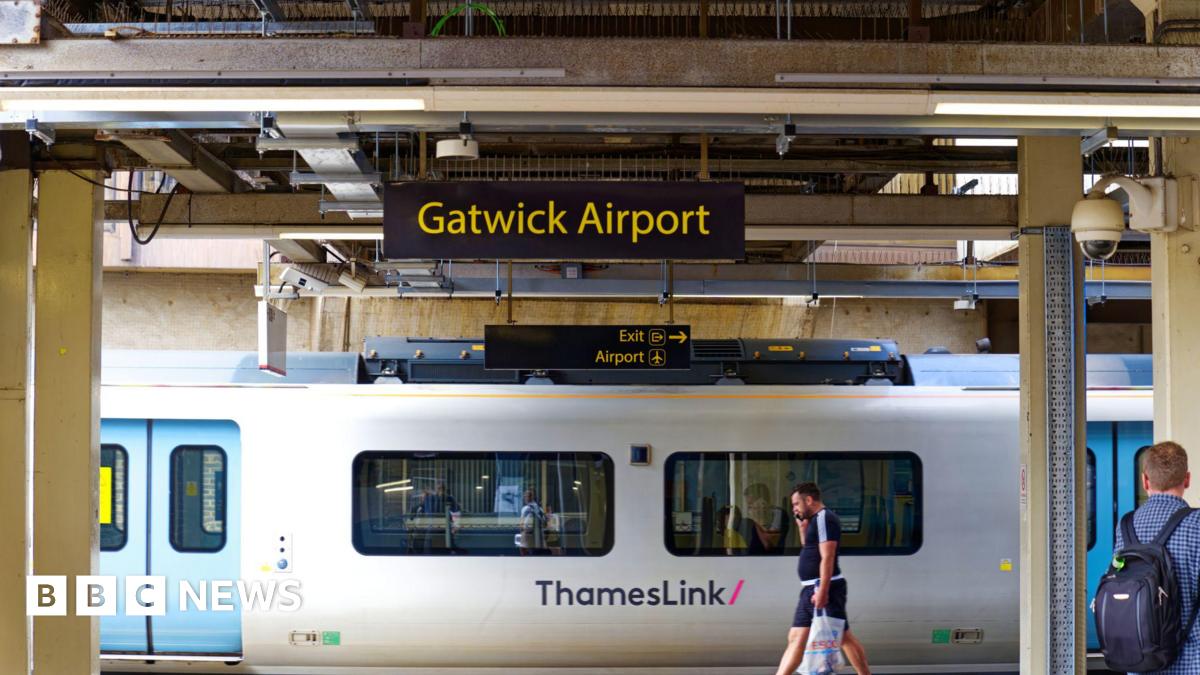 Sign at Gatwick Airport railway station with platform and a white Thameslink train departing the station. People are stood on the platform.
