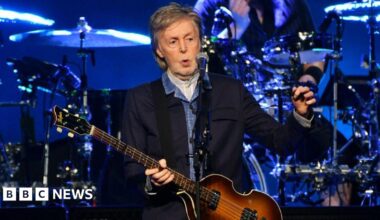 Sir Paul McCartney standing onstage. He has grey hair, and is wearing a dark blue jacket with light blue shirt. He is holding a bass guitar in his hands, with a drum kit visible in the background and a microphone in front of him.
