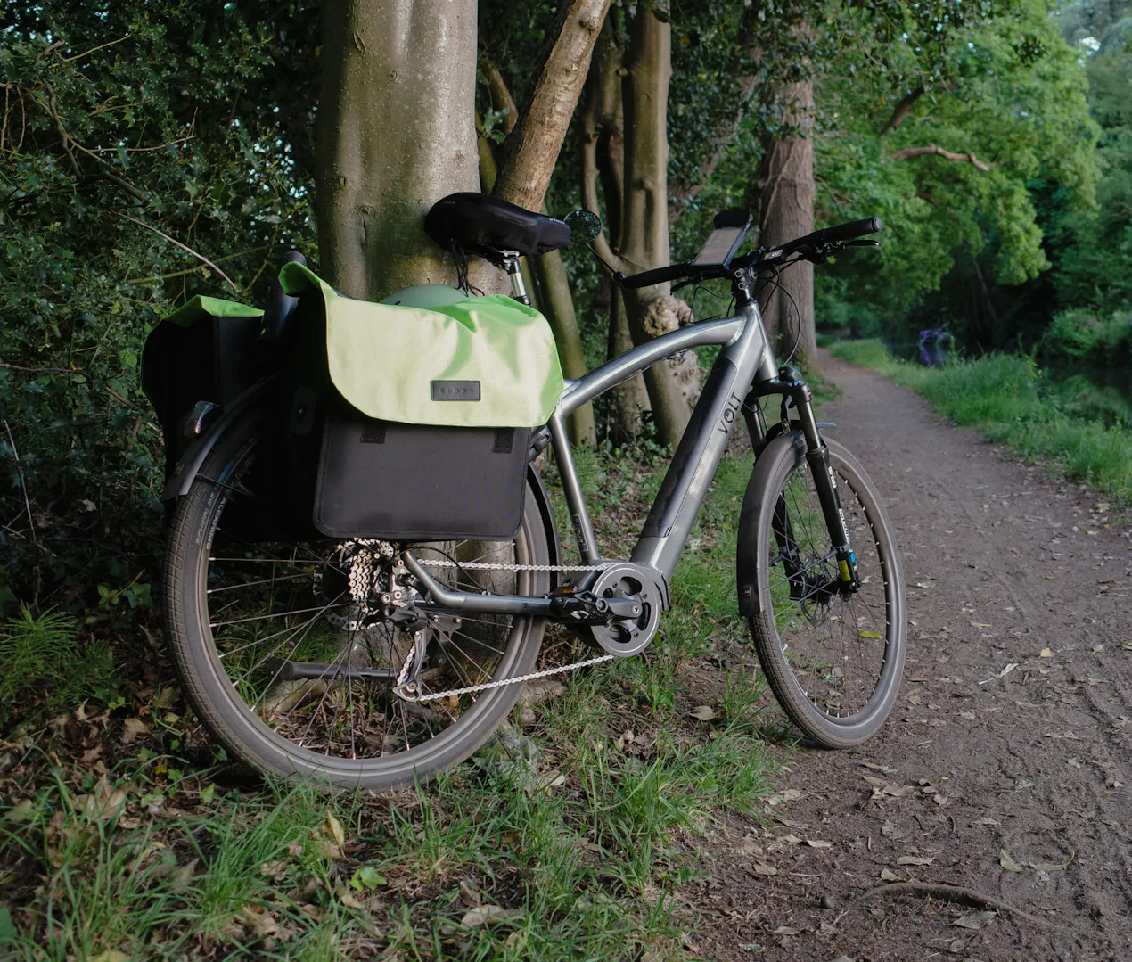 A bicycle with panniers resting against a tree on a dirt path next to a canal.