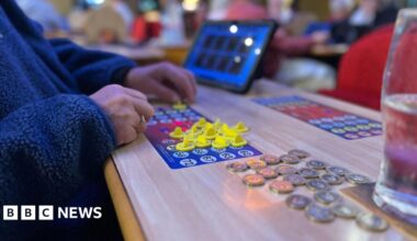A woman wearing a blue fleece moves bingo counters about on a board, with some pound coins on the table next to her.