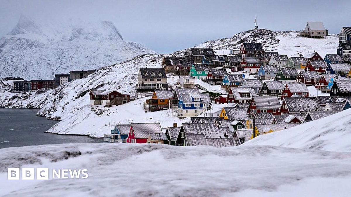 Houses of the old town are pictured in Nuuk, western Greenland. Around them are snowy rolling hills and a small mountain in the distance. The houses have snowy roofs, and ar different colours -- red, yellow, teal and blue, and are densly packed by the shore.