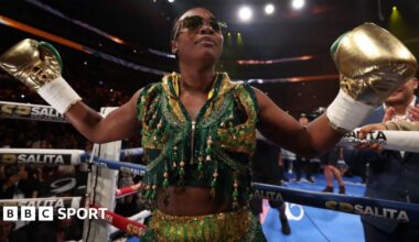 Claressa Shields holds out her arms and is wearing gold boxing gloves and a gold and green vest as she stands next to the ropes of a boxing ring