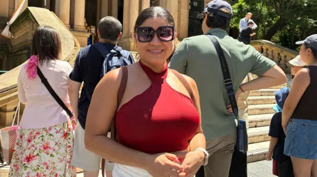 A woman in a red top and sunglasses smiling at the camera with a crowd in the background