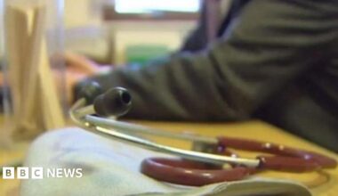 A stethoscope on a GP's desk with the doctor sitting at a yellow desk.