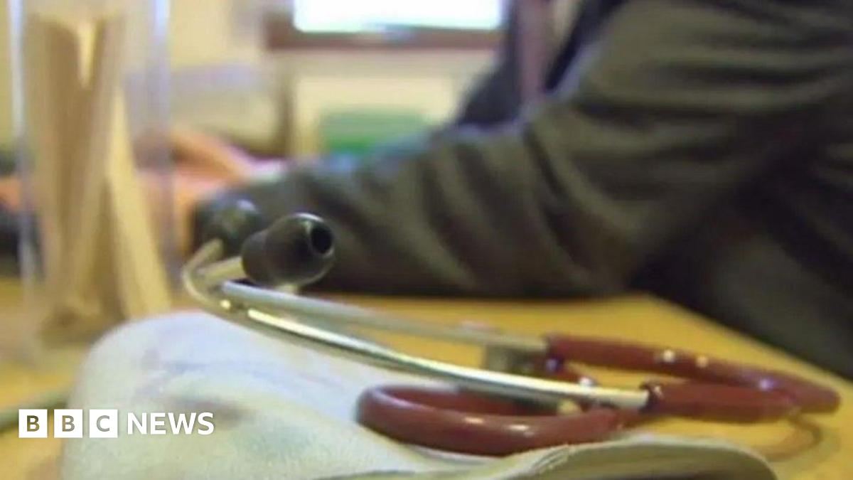 A stethoscope on a GP's desk with the doctor sitting at a yellow desk.