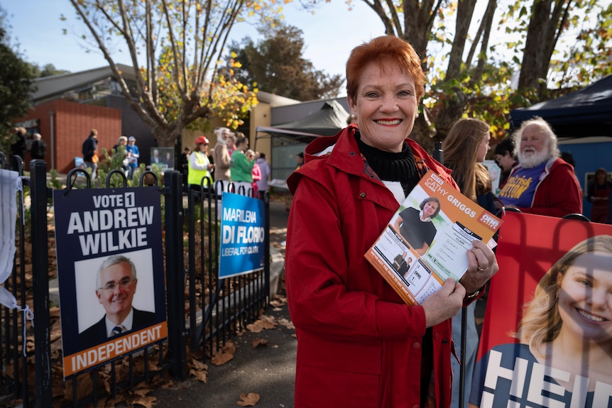 A woman with red hair, standing in front of election posters, holding flyers and smiling at the camera.