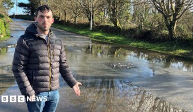 Luke Dunning is standing on a pavement and is pointing to the flooded road. There is a drain from which water is bubbling out. He is wearing a puffa jacket and has one headphone in.