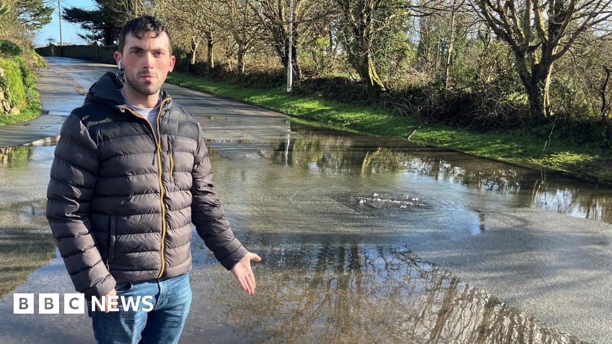 Luke Dunning is standing on a pavement and is pointing to the flooded road. There is a drain from which water is bubbling out. He is wearing a puffa jacket and has one headphone in.