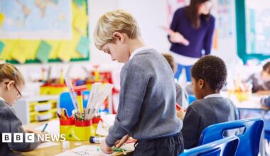 A blonde haired boy in a grey uniform stands at a desk doing a craft. Some other students are sitting at a table. A teacher at the top of the class wearing a purple top looks at some of their work.