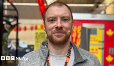 James is smiling at the camera. He is wearing a grey t-shirt and hoodie and has an orange lanyard around his neck.