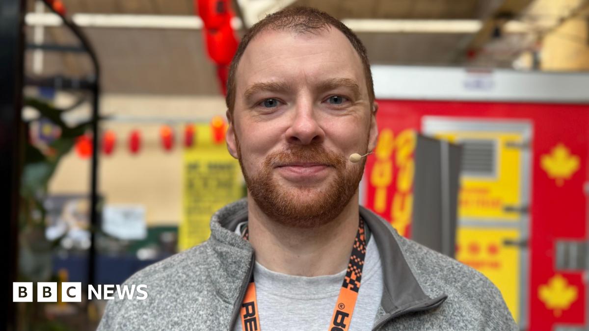 James is smiling at the camera. He is wearing a grey t-shirt and hoodie and has an orange lanyard around his neck.