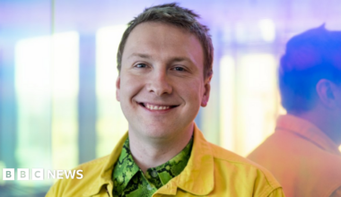 Joe Lycett pictured against a glass window. He is wearing a green snake print shirt underneath a yellow Demin jacket. He has short blond hair.