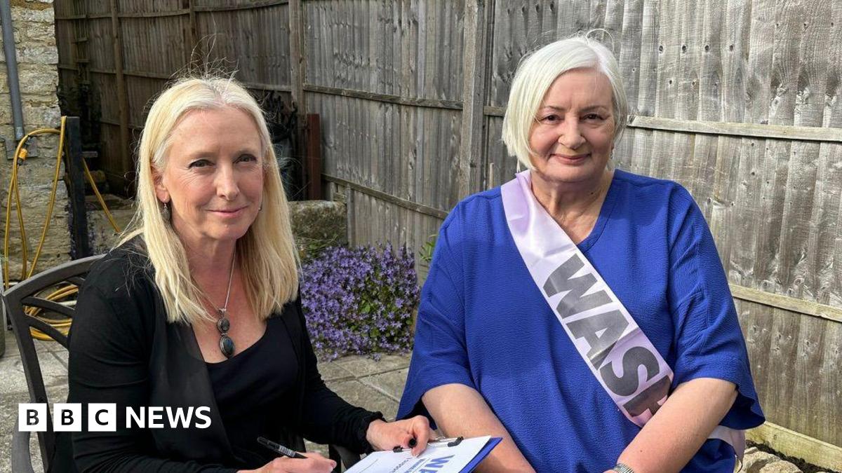 Two women are sat down on chairs in a garden. They are both smiling at the camera. One women has blonde hair and it wearing a black top and jacket. She is holding a clipboard with a piece of paper that has a large heading that reads 'Waspi'. The other women has short grey hair and is wearing a blue top. She is wearing a pink sash that says 'Waspi' on it.
