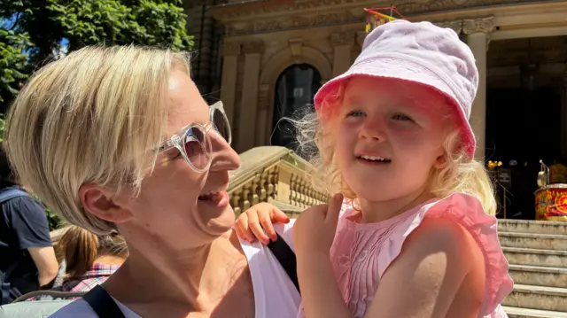 A woman with short blonde hair carrying a small child in a pink hat