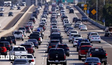 A freeway crowded with lines of cars, many with headlamps on, all bathed in the orange light of early morning in Los Angeles