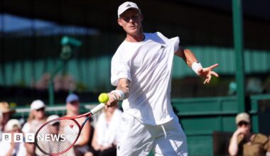 Billy Harris, who is dressed in white, swings a tennis racket at a yellow tennis ball at Wimbledon as the crowd looks on.