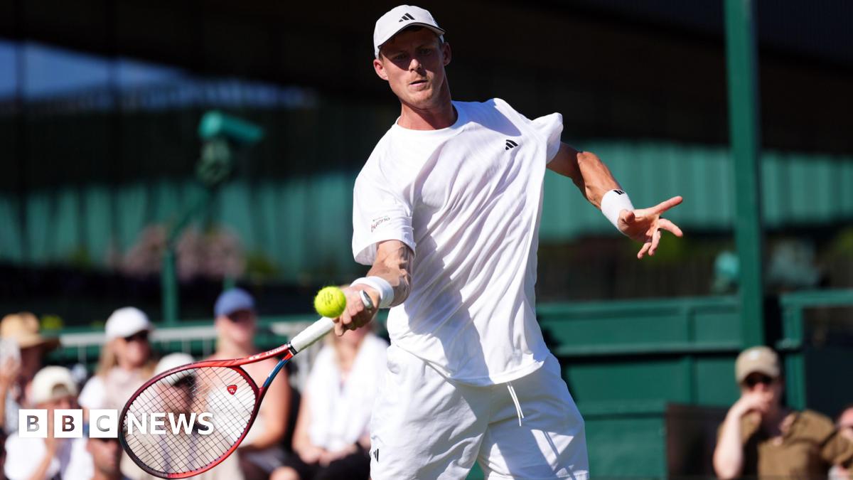 Billy Harris, who is dressed in white, swings a tennis racket at a yellow tennis ball at Wimbledon as the crowd looks on.