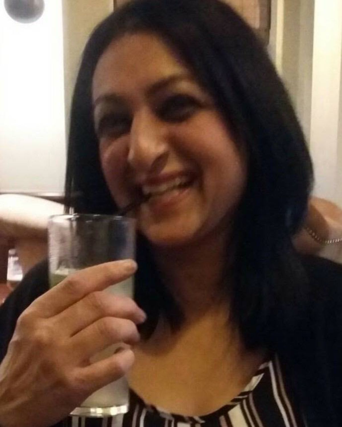 An individual with long, dark hair smiles brightly while holding a glass in a casual indoor setting.