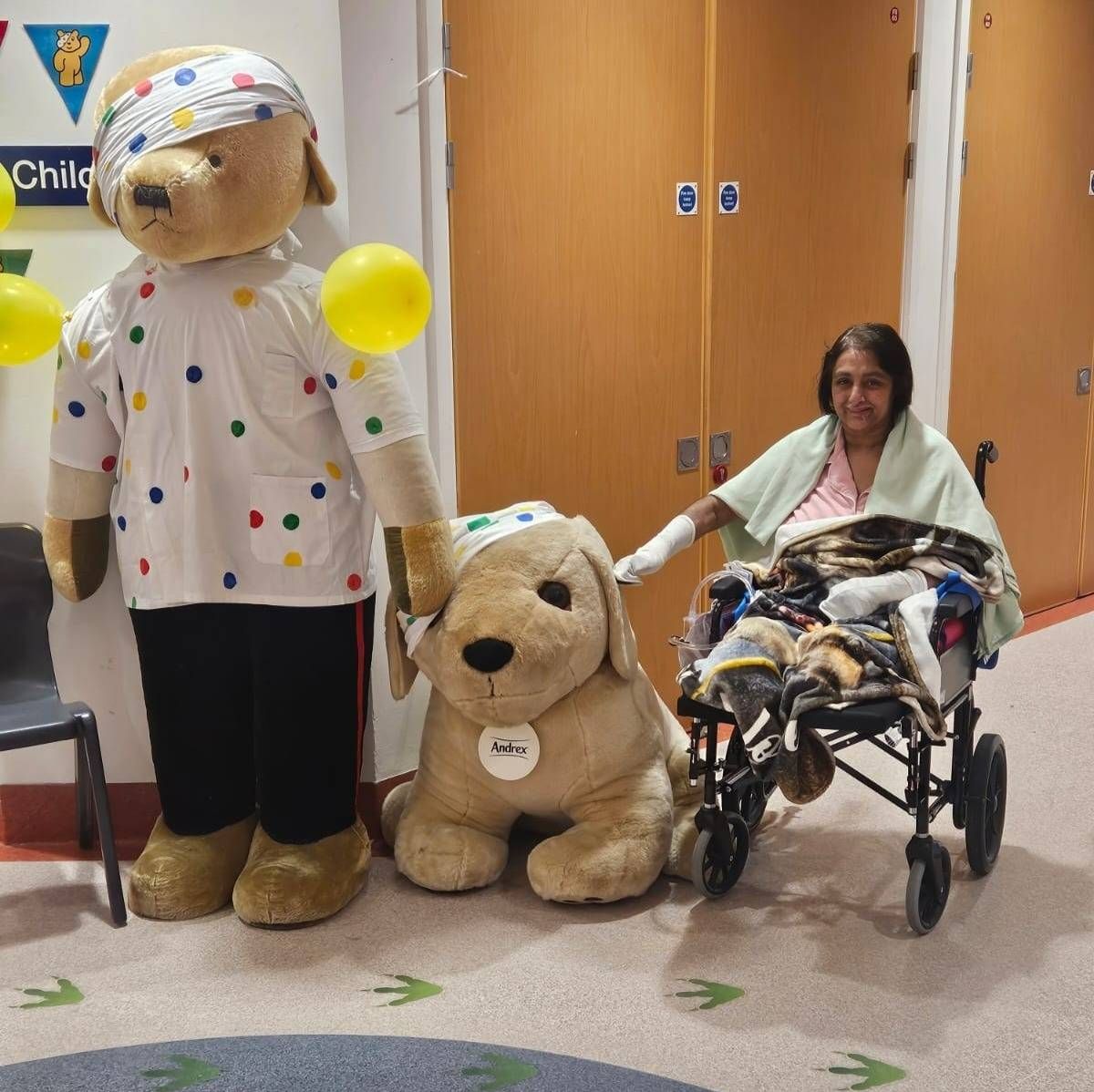 An individual in a hospital setting is being visited by a person dressed in a dog costume, accompanied by a life-sized dog plush toy, all of whom are holding yellow balloons.