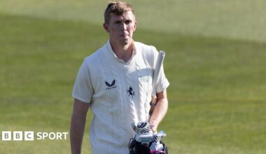 Zak Crawley walks off the cricket ground, dressed in a white short-sleeved top and white knitted vest, carrying a blue helmet and white and black gloves