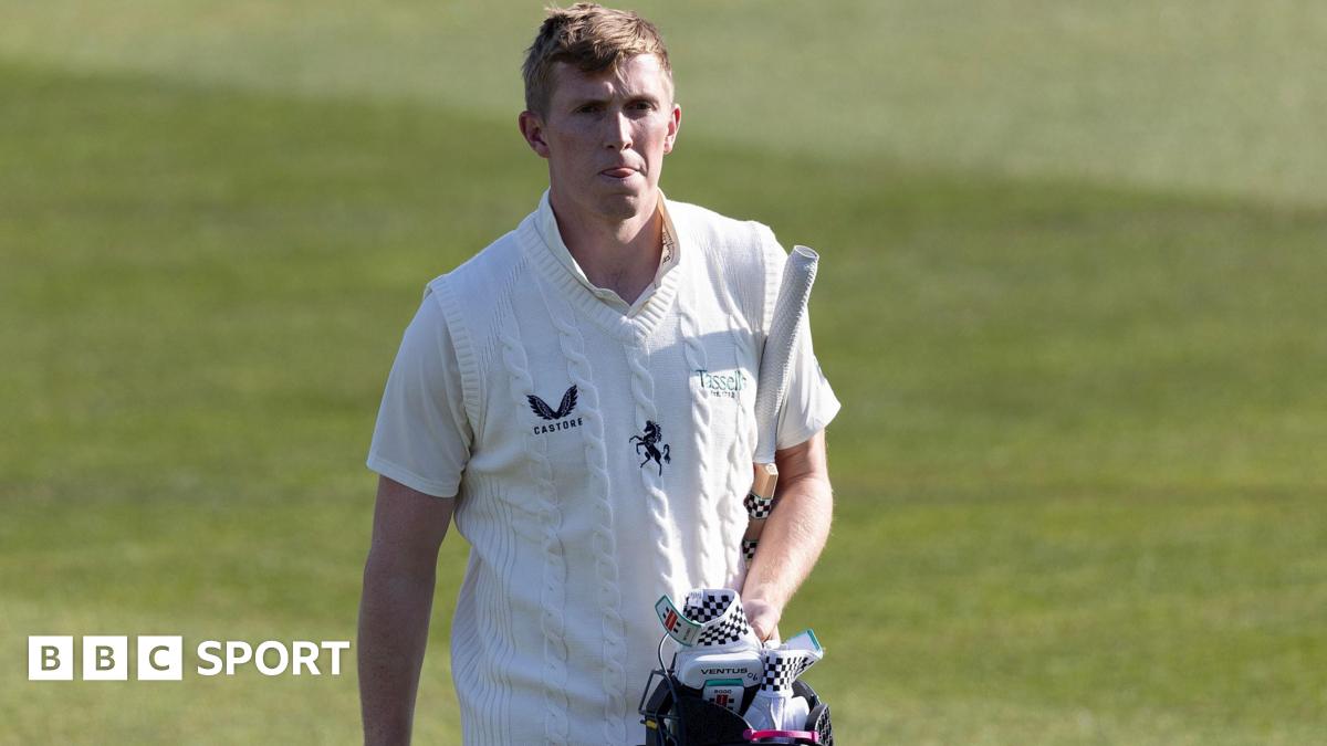 Zak Crawley walks off the cricket ground, dressed in a white short-sleeved top and white knitted vest, carrying a blue helmet and white and black gloves