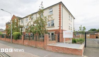 Exterior of a large three-storey building built of brick and partially covered in white rendering. It has black metal gates to the side, leading on to a car park, and a front garden filled with trees and plants