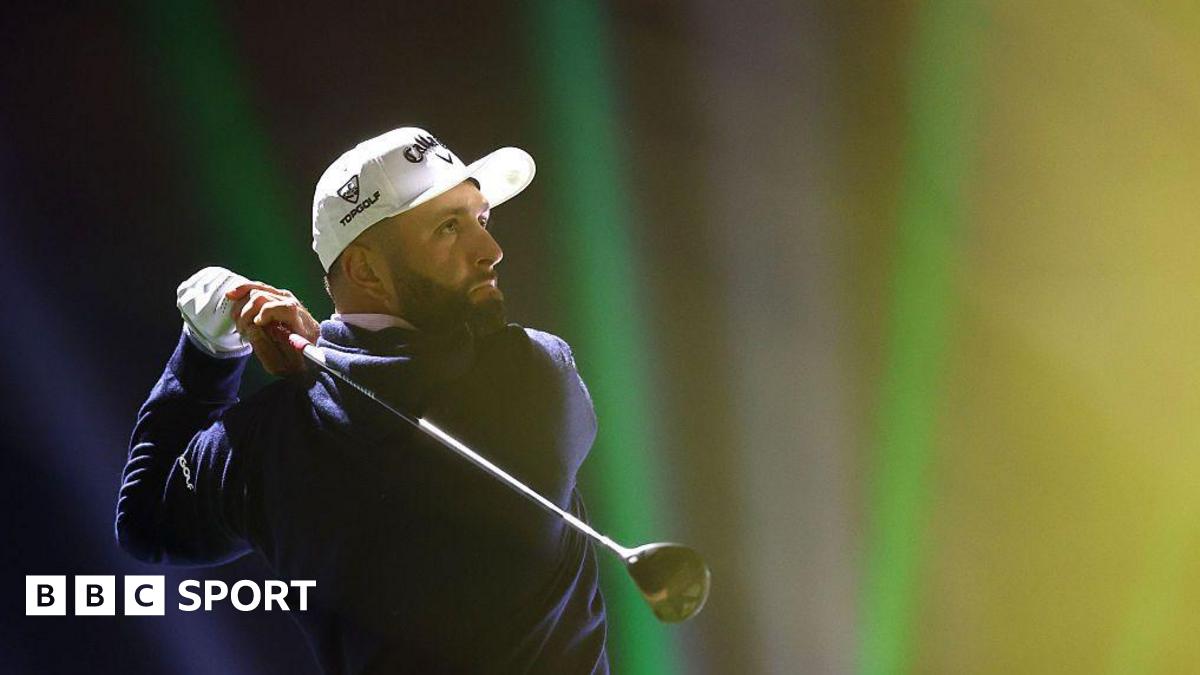 Jon Rahm tees off on the 16th hole on day one of LIV Golf Riyadh. He has a black beard and is wearing a white cap, a white glove on his left hand and a navy sweater.