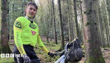 A man is stood in a forest next to a mountain bike. He is wearing a bright yellow jersey, black trousers and black and white gloves. He has brown hair in a mullet style. He is looking at the camera with a slight smile.