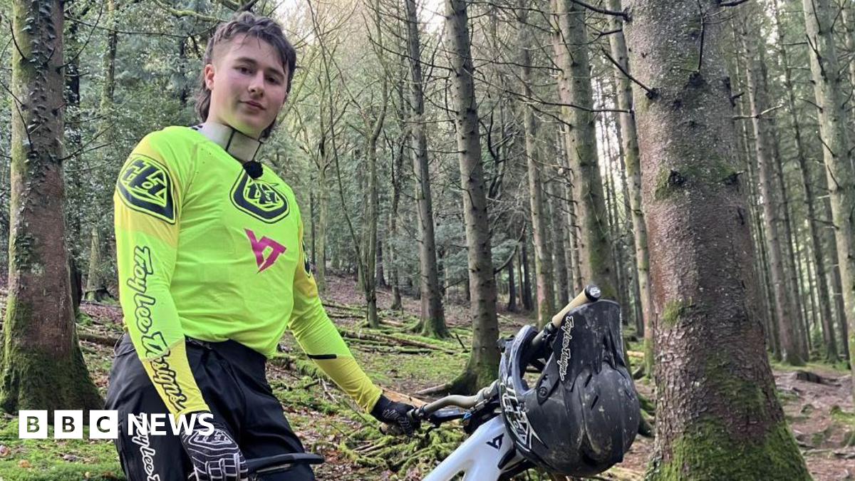 A man is stood in a forest next to a mountain bike. He is wearing a bright yellow jersey, black trousers and black and white gloves. He has brown hair in a mullet style. He is looking at the camera with a slight smile.