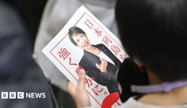 A person with their back to the camera holds a poster of Sanae Takaichi, wearing a black suit jacket, white t-shirt and pearl necklace. She is smiling. The poster has a white background and red border with red writing.
