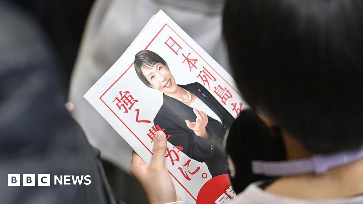 A person with their back to the camera holds a poster of Sanae Takaichi, wearing a black suit jacket, white t-shirt and pearl necklace. She is smiling. The poster has a white background and red border with red writing.